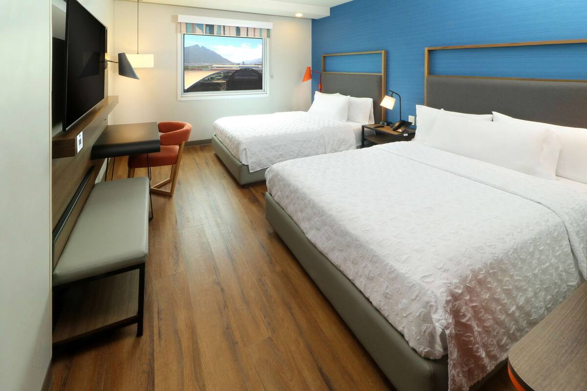 Quarto de hotel com duas camas, uma escrivaninha com cadeira, televisão e janela com vista para as montanhas.