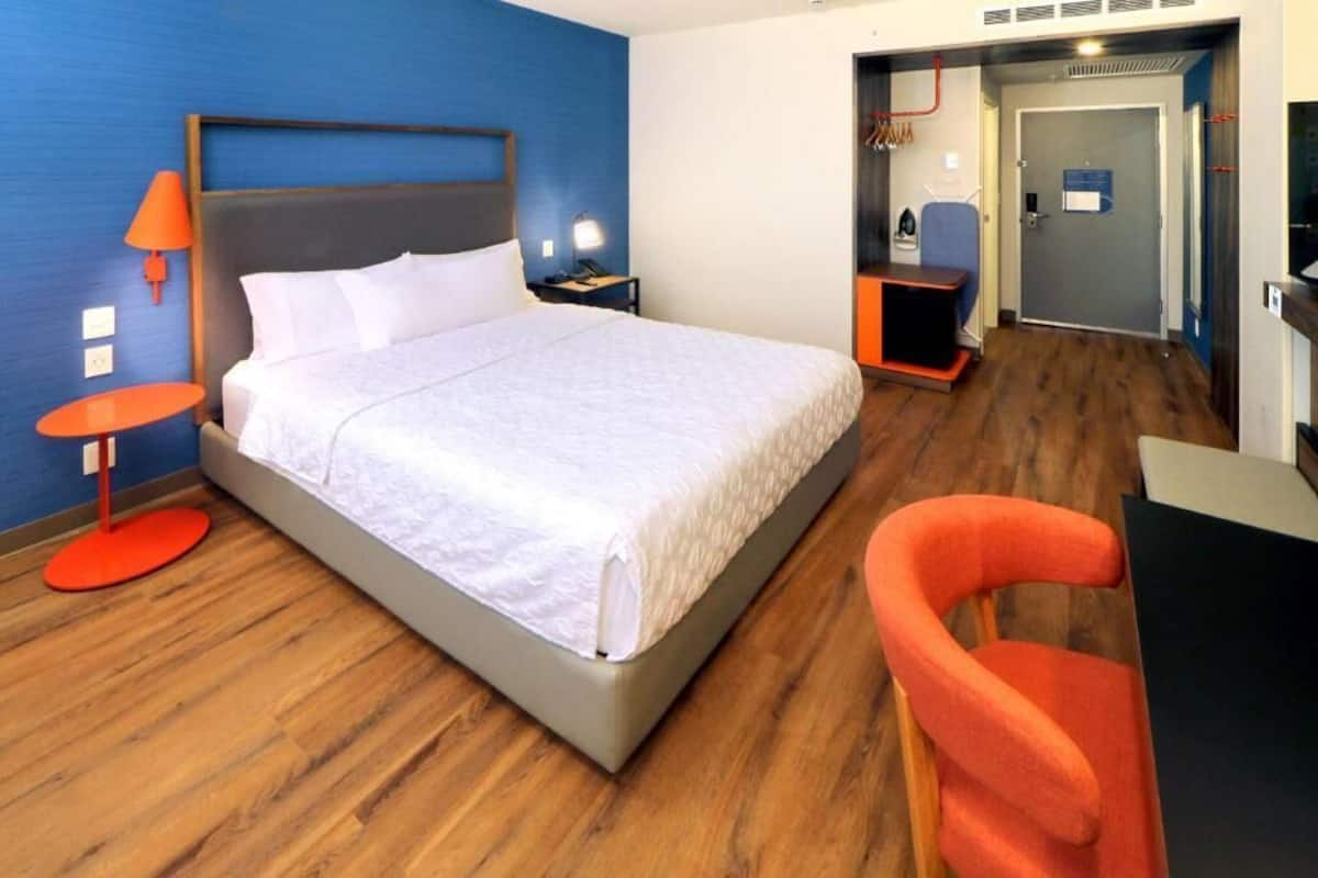 Quarto de hotel com cama, mesa de cabeceira, escrivaninha, cadeira e porta para o banheiro.