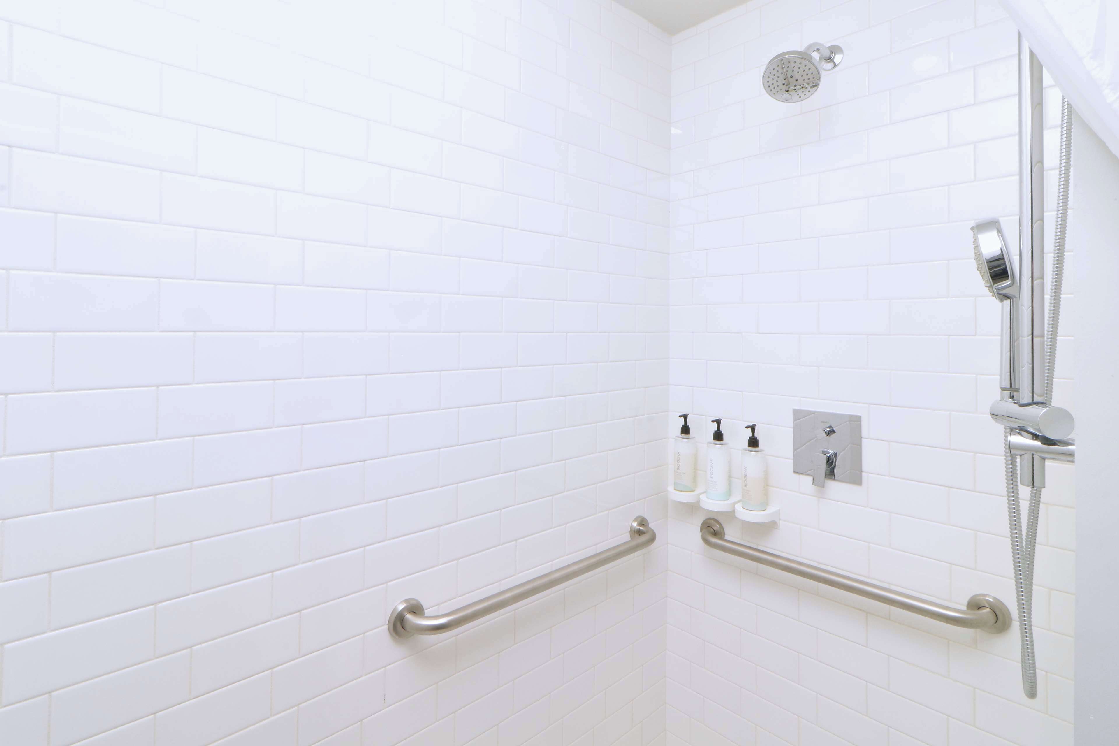 A white-tiled shower area with grab bars, a handheld showerhead, and a wall-mounted faucet.