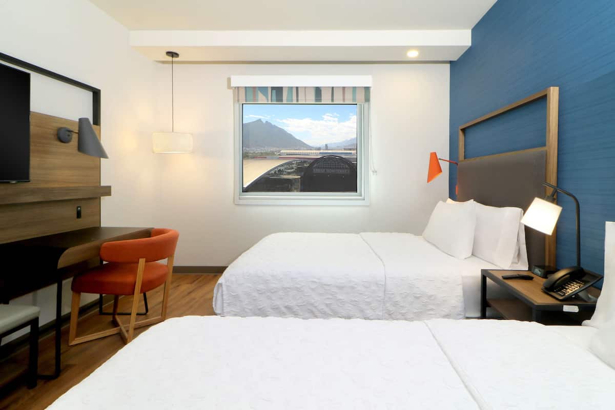 Quarto de hotel com duas camas, uma escrivaninha, uma cadeira, uma televisão, uma janela com vista para a montanha e uma mesa de cabeceira com abajur e telefone.