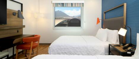 Habitación de hotel con dos camas, un escritorio, una silla, un televisor, una ventana con vistas a la montaña y una mesita de noche con lámpara y teléfono.