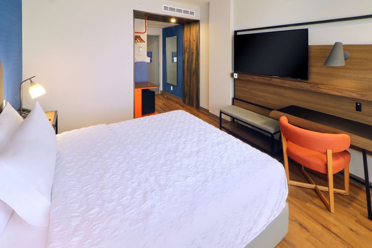 Quarto de hotel com uma cama grande, uma escrivaninha com cadeira, uma televisão na parede e um espelho.