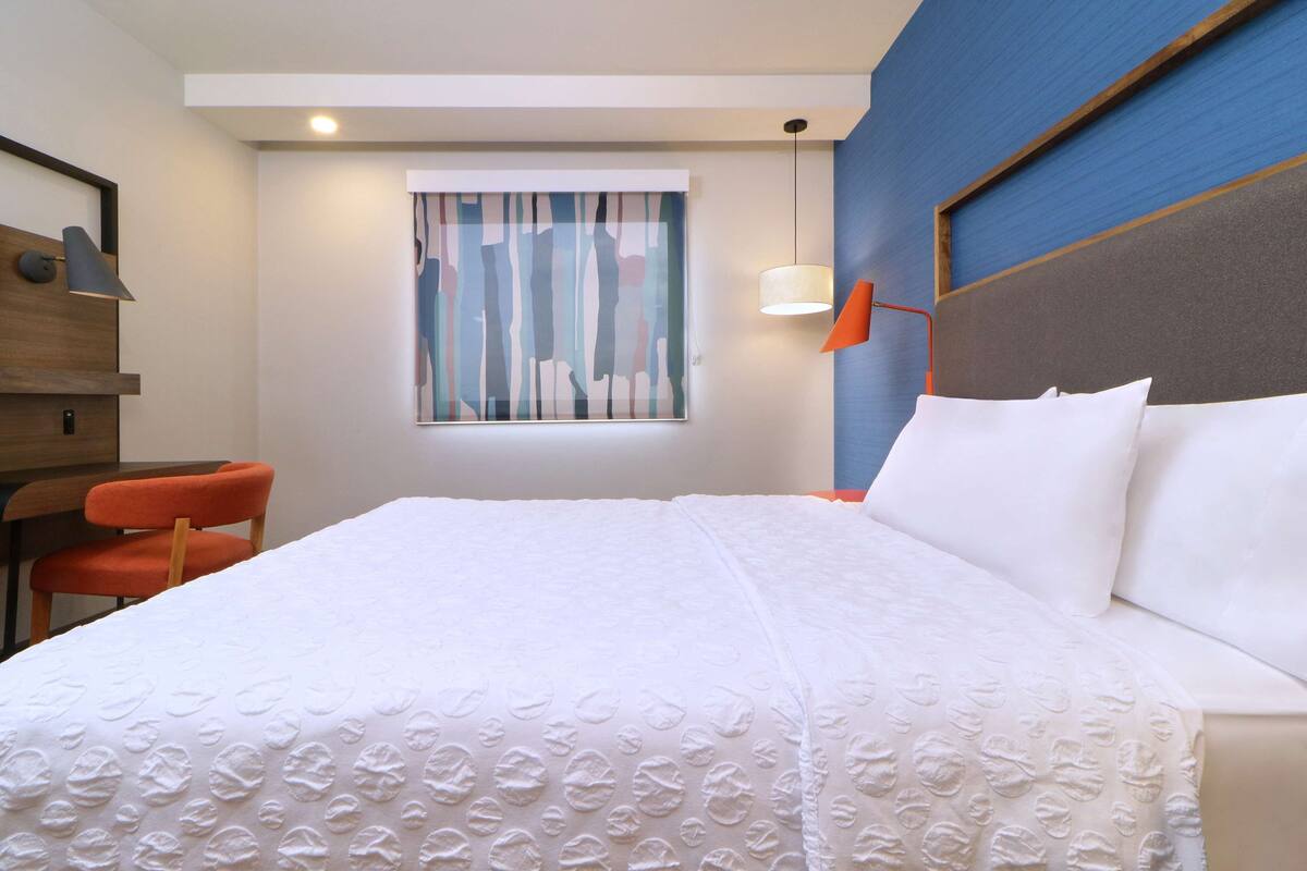 Quarto de hotel com cama, escrivaninha, cadeira, janela com cortinas e luminária fixada na parede.