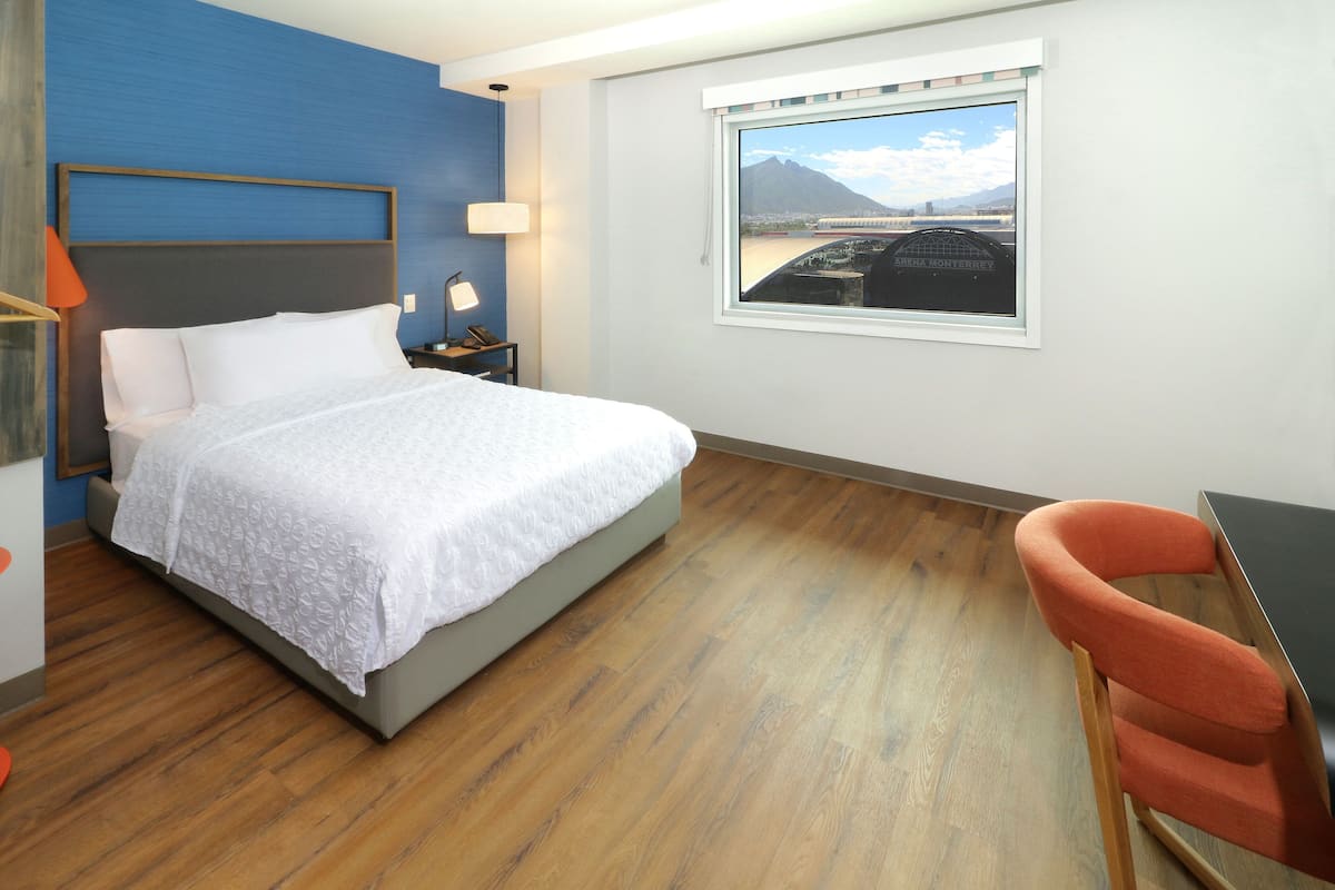 Quarto de hotel com cama, escrivaninha, cadeira, janela com vista para uma ponte e luminária.