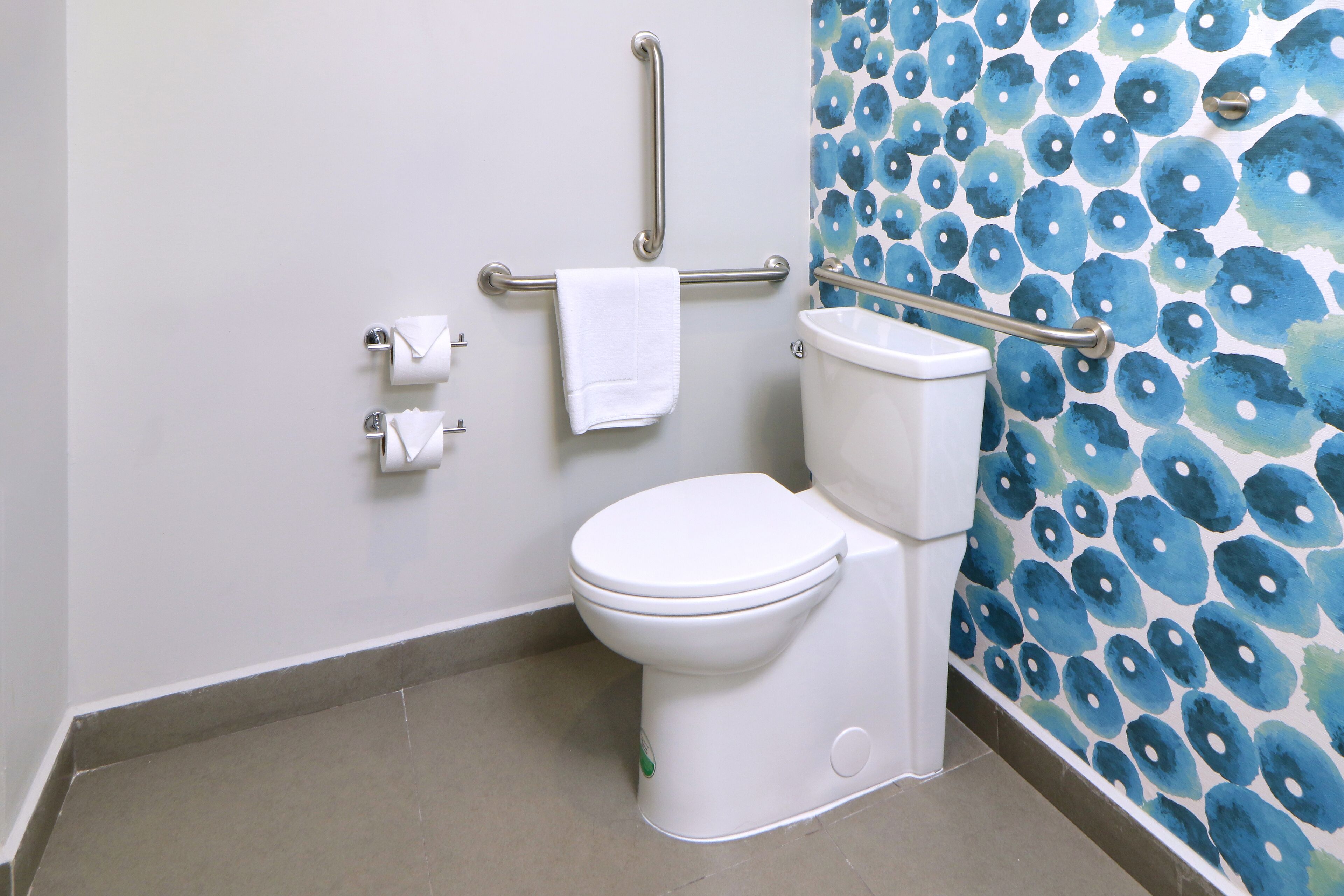 Un baño con inodoro blanco, un pasamanos y una pared con estampado floral azul.