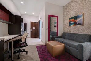 A modern living room with a grey sofa, a small wooden table, and a chair. There is a large painting on the wall and a door with a mirror. - Home2 Suites By Hilton Redding (Redding)