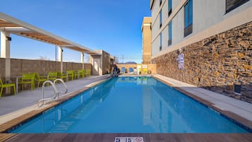 A swimming pool with a stone wall on one side and a covered patio area with chairs on the other.