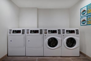 A row of four white commercial laundry machines in a room with plain walls and a wooden floor. - Home2 Suites By Hilton Redding (Redding)
