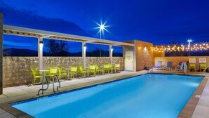 A swimming pool with chairs and tables, a covered area with lights, and a stone wall. - Home2 Suites By Hilton Redding (Redding)