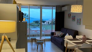 A modern living room with a sofa, dining table, and chairs. There is a large window offering a view of the outdoors.