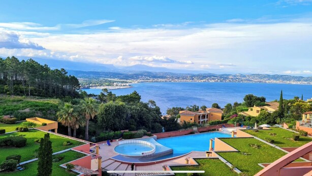 A resort with a large swimming pool, lush greenery, and a view of the sea and distant cityscape.