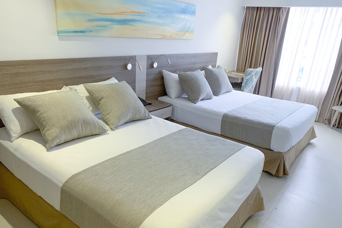 A hotel room with two beds, a nightstand, a lamp, a painting, and a window with curtains.