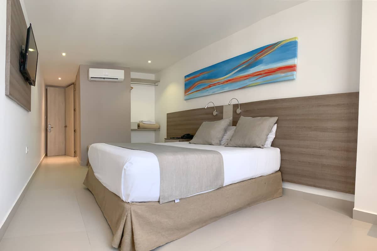 A modern hotel room with a large bed, a flat-screen TV mounted on the wall, an air conditioning unit, and a colorful abstract painting above the bed.
