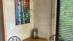A corner room with wooden walls, a round wooden table, two upholstered chairs, a metal sculpture, and a window with blinds. - The Riverbirch Treehouse-close to town yet steps away from total wilderness (Nixa)