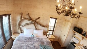 A cozy bedroom with a large bed, a wooden headboard, a chandelier, and a small kitchenette. - The Riverbirch Treehouse-close to town yet steps away from total wilderness (Nixa)