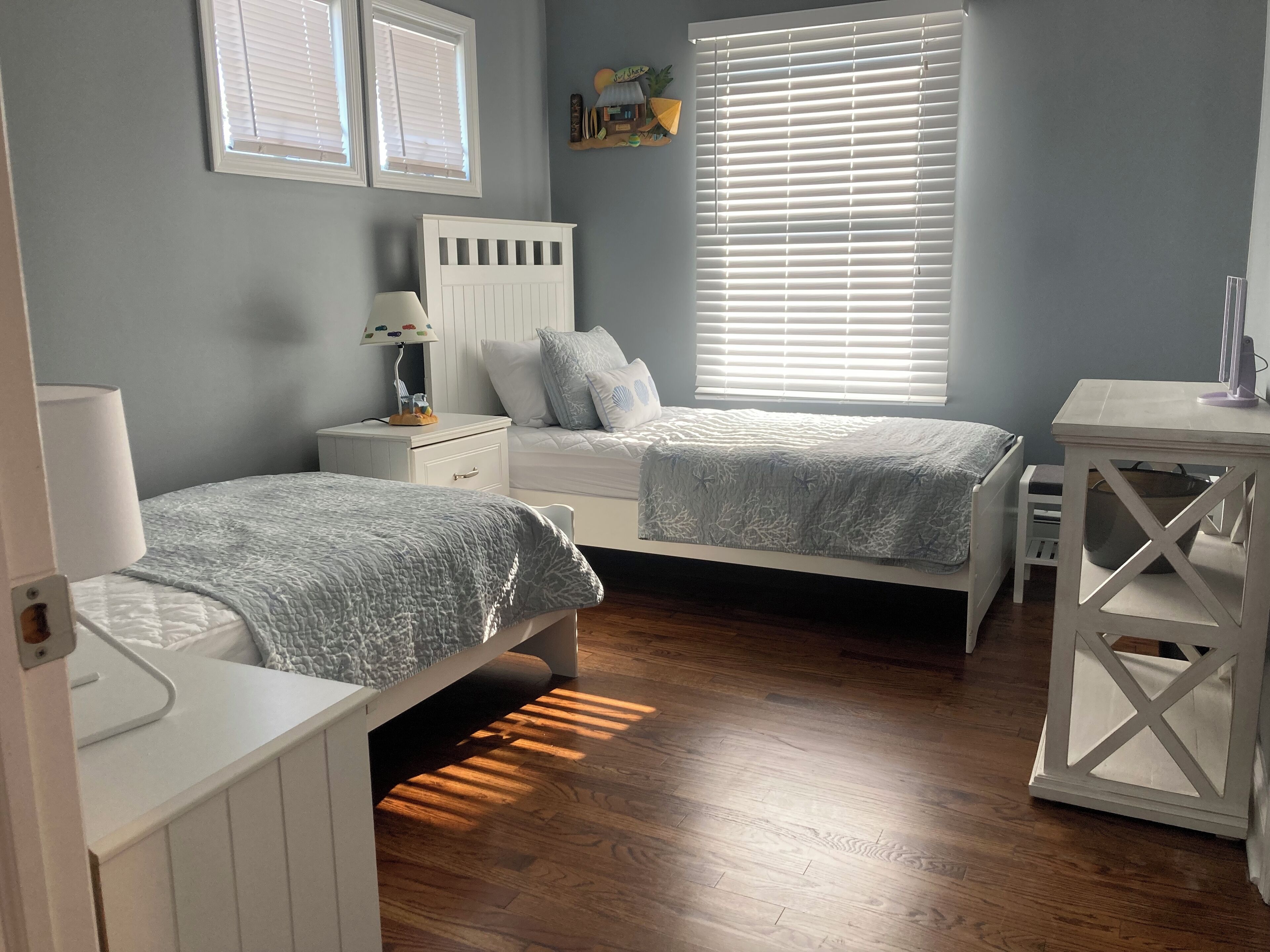 A bedroom with two single beds, a nightstand with a lamp, a dresser, a window with blinds, and a small table with a lamp.