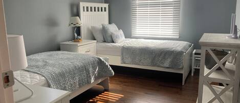 A bedroom with two single beds, a nightstand with a lamp, a dresser, a window with blinds, and a small table with a lamp.