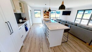 Una cocina moderna con armarios blancos, una isla central, iluminación colgante y un sofá gris en un espacio luminoso con ventanas amplias.