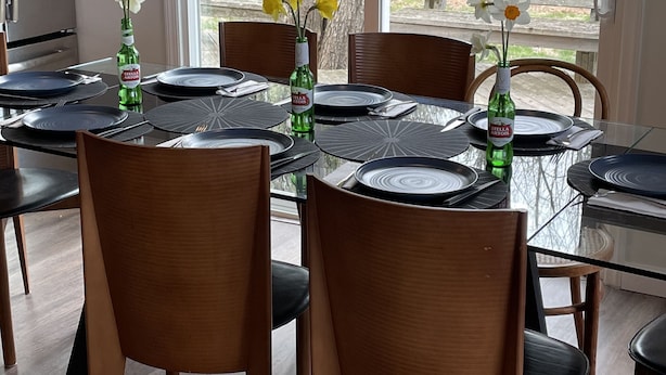 Un comedor con una mesa de vidrio rodeada de sillas de madera, un refrigerador y una ventana con vista a árboles.