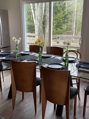 A dining area with a glass table surrounded by wooden chairs, a refrigerator, and a window with a view of trees.