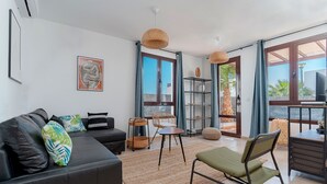 A modern living room with a sofa, a dining table, and a chair. There is a large window with curtains, a wicker chair, and a small table with a basket. - 7 PIEDRAS (Playa Blanca)