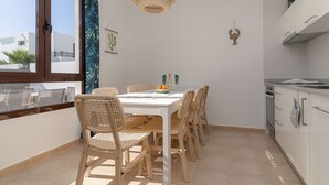 A compact kitchen with a dining area, featuring a white table, wicker chairs, a hanging light fixture, and a view through a window. - 7 PIEDRAS (Playa Blanca)