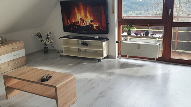 Ein modernes Wohnzimmer mit einem Flachbildfernseher, der einen Kamin zeigt, einer Couch, einem Couchtisch und einer Aussicht auf eine Berglandschaft durch große Fenster.