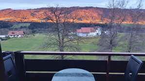 Ein Balkon mit Tisch und zwei Stühlen, mit Blick auf eine ländliche Landschaft mit Häusern und Bäumen.