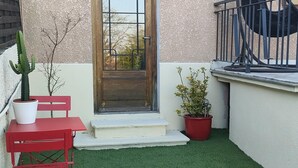 Eine kleine Terrasse mit Kunstgrashalm, einer Holztür mit Metallgitter, einem roten Tisch und Stuhl aus Metall sowie zwei Blumentöpfen.