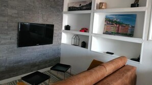 Ein modernes Wohnzimmer mit einem flachbildfernseher an einer Ziegelwand, einem braunen Ledersofa und weißen Regaleinheiten mit Dekorationselementen.