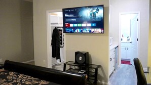 Un dormitorio con una cama, un televisor montado en la pared, un altavoz sobre una mesita y un armario con ropa colgada.