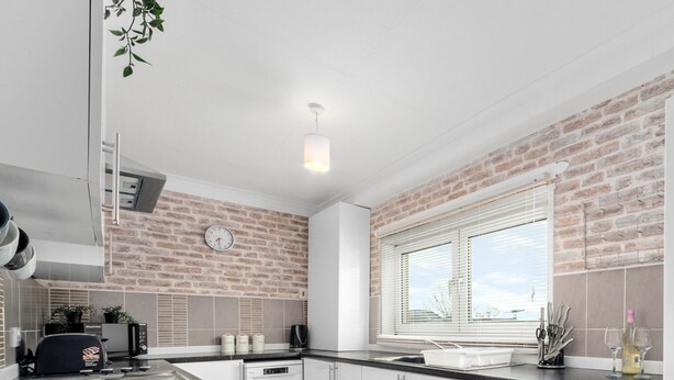A modern kitchen with white cabinets, a brick accent wall, a built-in washing machine, and a window with blinds. - Three Bedroom Apartment by Klass Living Serviced Accommodation Coatbridge - Berwick Apartment With WiFi &Parking (Coatbridge)