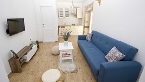 A compact living space with a blue sofa, a flat-screen TV mounted on the wall, a small white table with a vase, and a kitchen area visible through an open door. - Cozy, modern,new apartments - 1 minute from the beach (Drašnice)