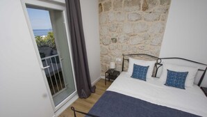 A bedroom with a stone wall, a balcony with a view, a bed with blue and white bedding, and a small bedside table with a lamp. - Cozy, modern,new apartments - 1 minute from the beach (Drašnice)