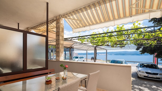 En balkon med et glasbord, fletstole og udsigt til havet og parkerede biler.