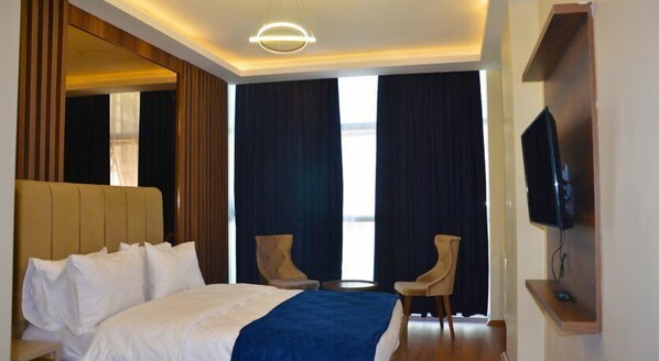 A hotel room with a large bed, two chairs, a TV, and a large window with curtains.