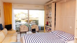 A room with a bed, a sofa, a TV, a laptop, two chairs, a bookshelf, and a sliding glass door leading to an outdoor area with a table and chairs. - Lovely sea view studio in Locmariaquer (Locmariaquer)
