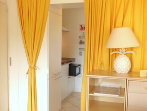 A room with yellow curtains, a white lamp shaped like a pineapple, and a kitchen area visible through an open door. - Lovely sea view studio in Locmariaquer (Locmariaquer)