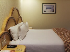 Habitación de hotel con cama, almohadas, mesita de noche con control remoto, un cuadro en la pared y una manta estampada.