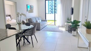A modern living room with a grey sofa, a dining table with chairs, a TV mounted on the wall, and a large window with a view of a building. - Luxury apartment in Doral miami, 1 Bd (Doral)