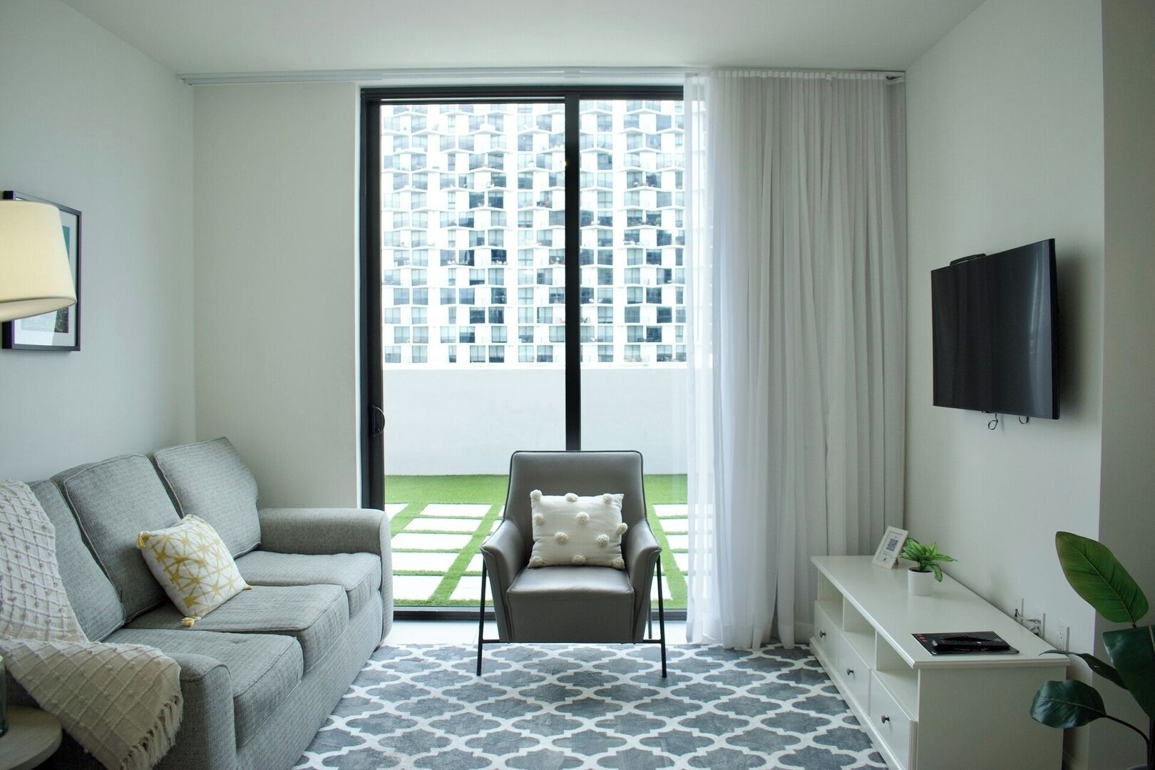 A modern living room with a grey sofa, a grey armchair, a flat-screen TV, and a view of a high-rise building through a glass door.