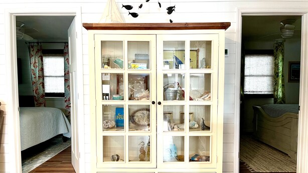 Une pièce avec un meuble blanc aux portes vitrées, un plancher en bois et un lit agrémenté de rideaux à motifs floraux.