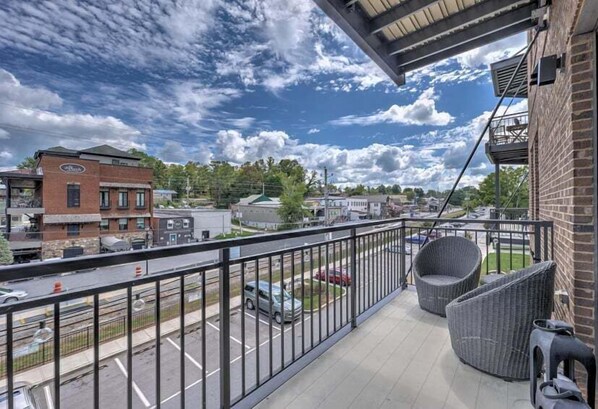 A balcony with wicker furniture, a view of a parking lot, and a brick building in the background. - Nest on West - Small town charm | Downtown Condo | Walk to shops (Blue Ridge)