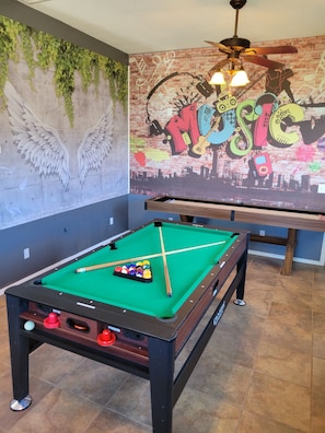 Ein Billardtisch mit Kugeln und Spielstöcken, ein Deckenventilator und ein Wandgemälde im Graffiti-Stil.