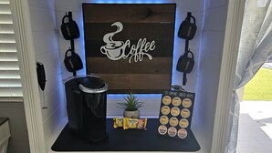 A coffee station with a sign reading "Coffee," a coffee maker, a plant, and a display of coffee pods. - Lodging and entertainment all wrapped into one great place! (Apache Junction)