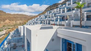 A row of white buildings with blue accents, likely part of a hotel or resort, with a mountainous backdrop. - Club Cala Blanca (MOGAN)