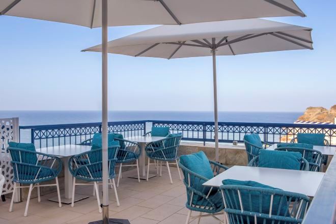 An outdoor seating area with tables and chairs, a white umbrella, and a view of the sea.
