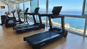 A gym with treadmills and exercise equipment, large windows with a view of the sea. - Club Cala Blanca (MOGAN)