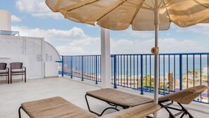 A balcony with two lounge chairs, a large umbrella, and a view of the sea and buildings. - Club Cala Blanca (MOGAN)