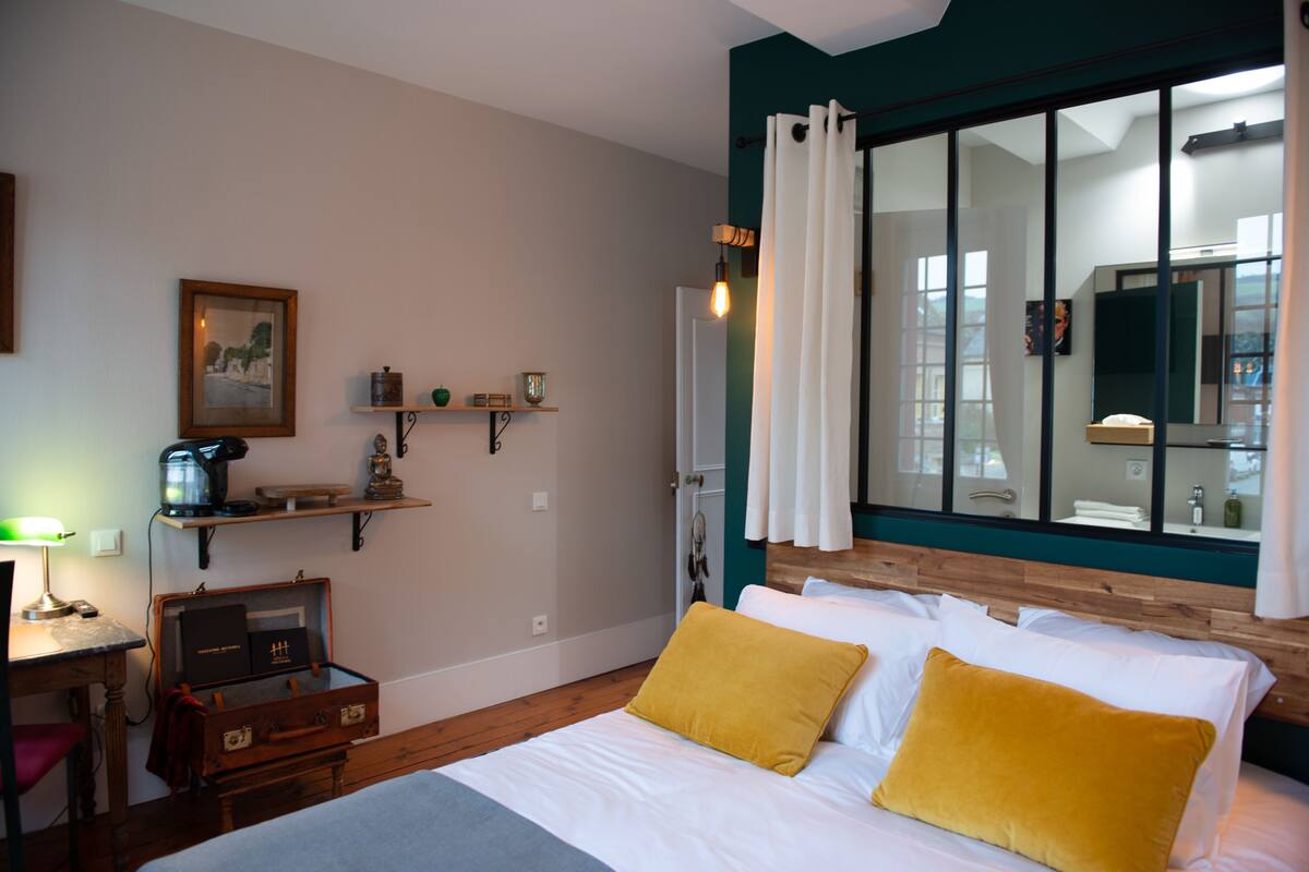 A hotel room with a bed, a desk, a chair, a suitcase, a shelf with decorative items, and a large window with curtains.